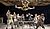 A performer leaps above a table surrounded by seven other actors, all dressed in period costumes, in a theatrical scene.