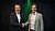 Two men in business attire shake hands, standing against a dark backdrop with green lighting.