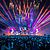 A large crowd at a concert enjoys a vibrant light show with colorful beams and fireworks illuminating the stage and audience.
