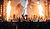 A live music performance featuring multiple musicians on stage, with dramatic lighting, flames, and a large screen displaying visuals in the background.